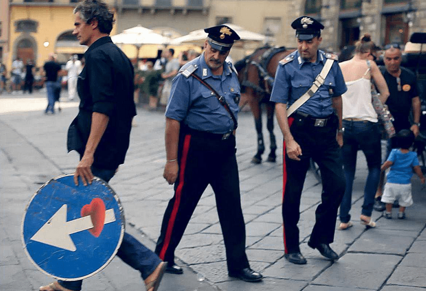 due carabinieri osservano perlessi clet che porta a spasso un segnale stradale modificato