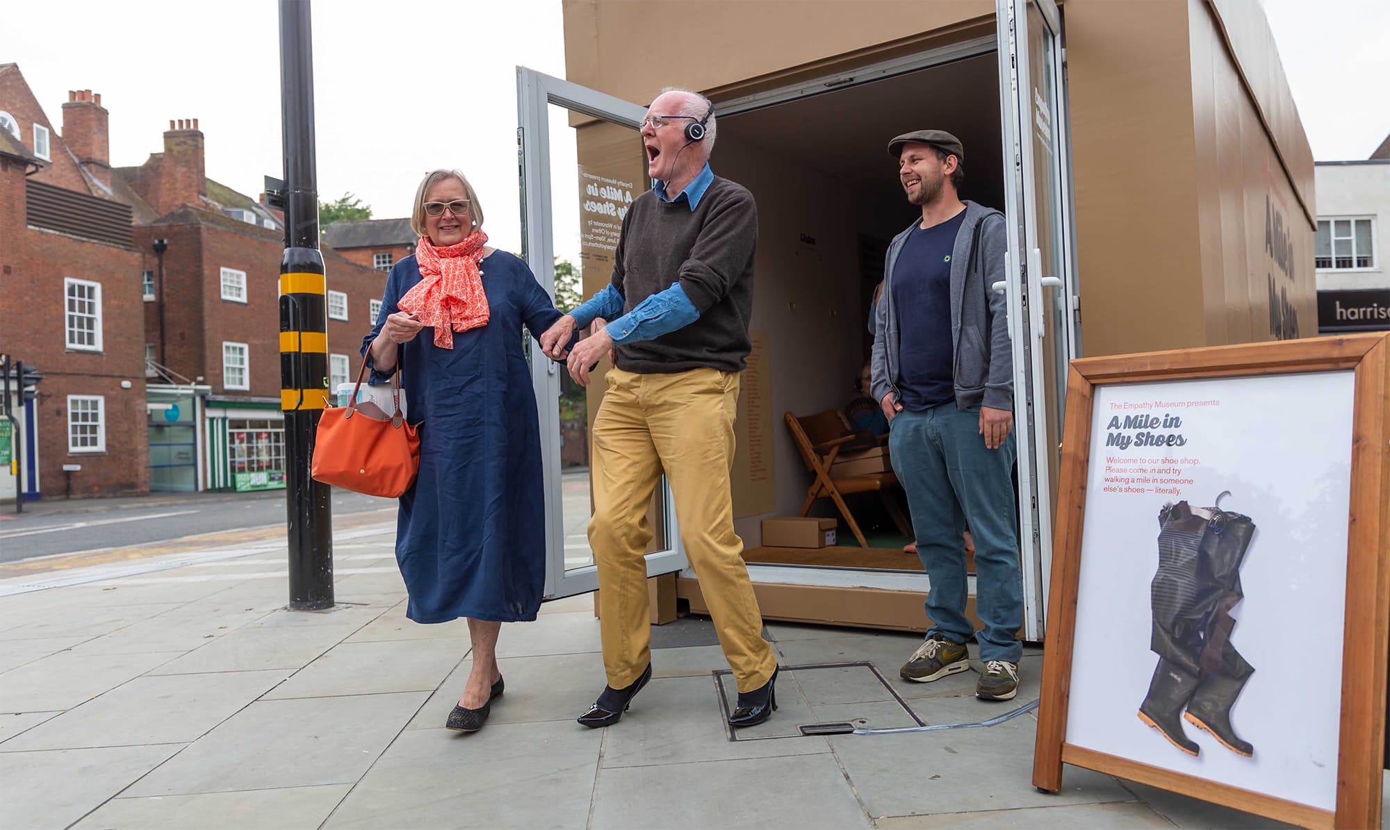 un signore anziano si sta divertendo a camminare su delle scarpe coi tacchi (forse della moglie che lo sta sostendendo per mano?) mentre un addetto di Empathy Museum osserva divertito